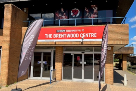 Image showing the entrance to the Brentwood Centre.  A red sign reads 'EVERYONE WELCOMES YOU TO THE BRENTWOOD CENTRE', with two sets of doors underneath.  Two grey 'Everyone Active' flags are placed by the entrance.