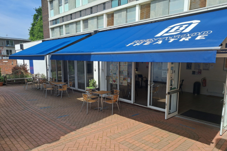 Image of the exterior of Brentwood Theatre, with blue awning, tables and chairs outside, and double doors open
