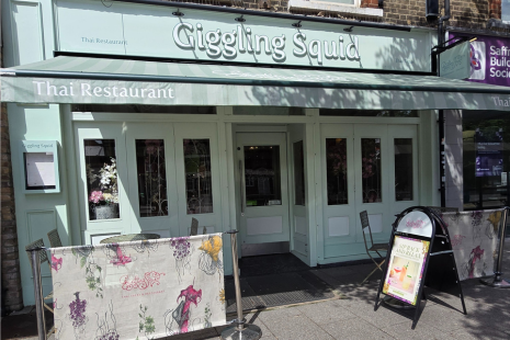 Image of the exterior of Giggling Squid in Brentwood, painted pale green with white signage.  A cream banner with flowers is outside, in front of a seating area.