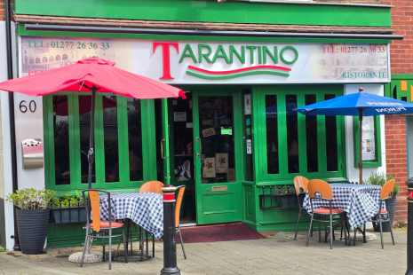 Image showing the exterior of Tarantino restauran.  The doors and windows are green, while the signage is green and red.  Two tables with blue gingham tablecloths and parasols are outside.