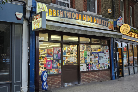 Image showing the exterior of Brentwood Mini Market, with yellow and blue lettering, a lottery sign and adverts placed in the windows.