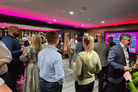 This image shows a group of people attending a business event in a dimly lit room with a modern, upscale ambience. The room features pinkish-purple LED lighting along the ceiling edges, and the walls display illuminated screens with bright colours. Attendees are dressed in business casual attire, standing in small groups and engaging in conversation. Most people have their backs or sides turned to the camera, and they appear to be socialising or networking. 