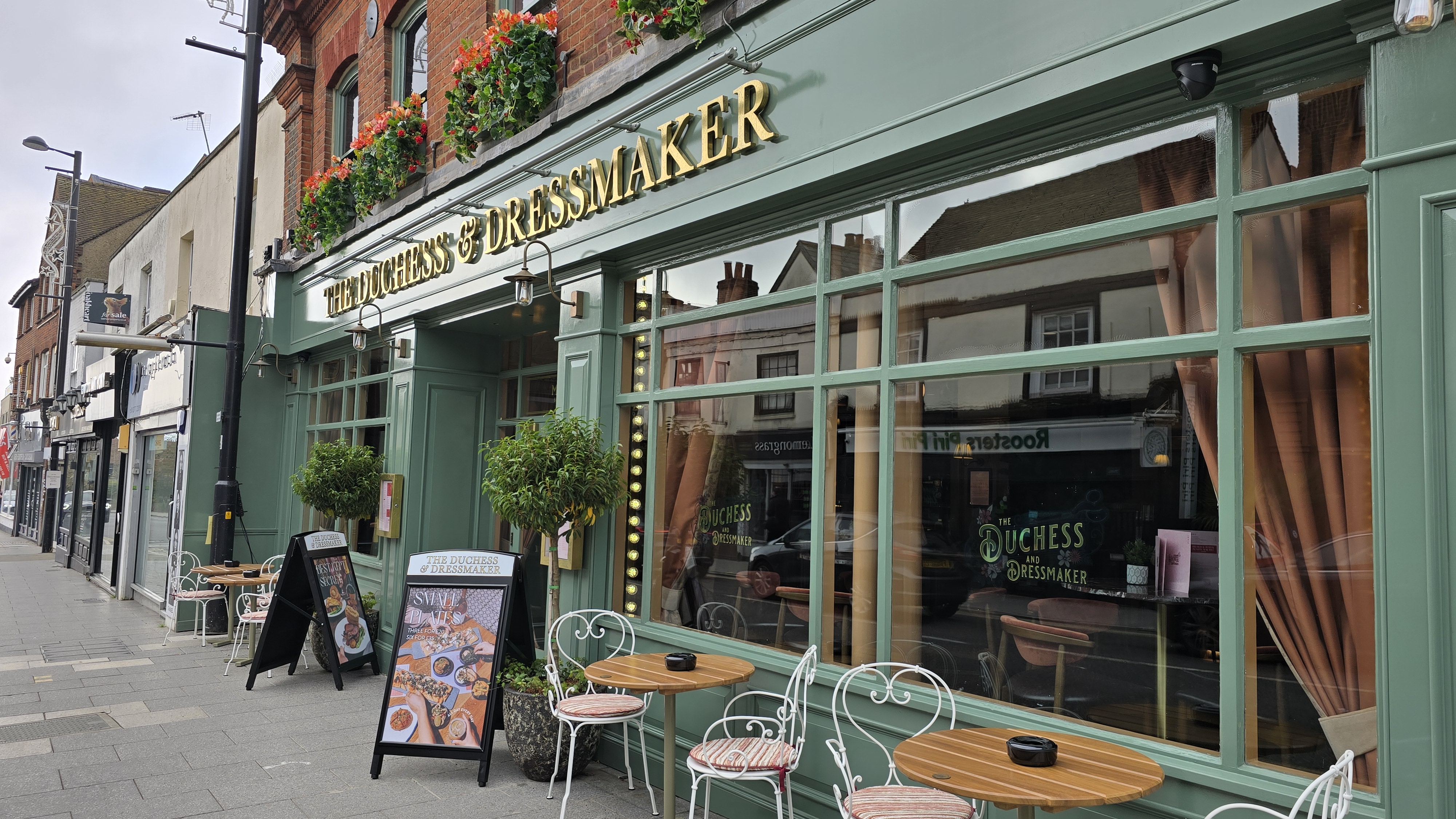 Image showing the exterior of The Duchess & Dressmaker, painted green with gold writing and velvet curtains at the windows.  Two tables and four chairs are outside, while window boxes with red flowers are visible outside the first floor windows.