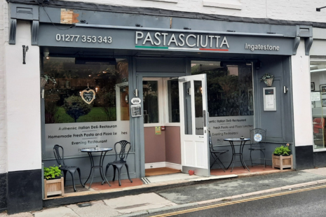 Image showing the exterior of PASTASCIUTTA restaurant in Ingatestone, with grey signage and white lettering, with a green, white and red line underneath, reminiscent of the Italian flag.  On either side of the door is a table and two chairs.