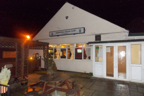 Image showing the exterior of the Bardswell Social Club.  There is outdoor seating and warm lights.