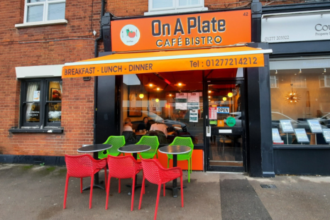 Image showing the exterior of On A Plate in Shenfield.  There are red and green chairs and tables outside, while the signage and awning is orange.