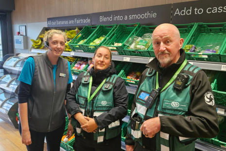 Town Rangers Andrew and Chelsie, wearing green Brentwood Connected tactical vests, standing in front of green racks of fruit and vegetable products and a member of staff in Co-Op in Shenfield