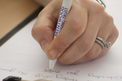 An image of a hand, wearing two rings, writing on a piece of paper with a pen which reads 'BRENTWOOD HOUSING TRUST'.