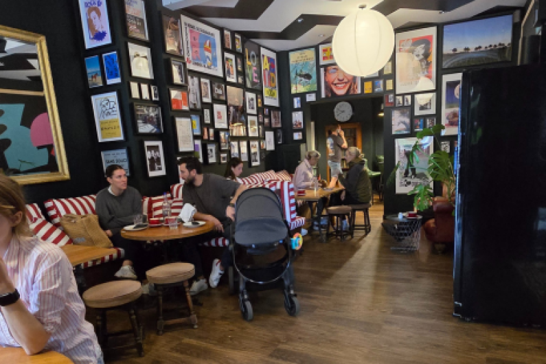 The image shows a cosy café or lounge area with multiple people seated at round wooden tables. Two people on the left are engaged in conversation, while a buggy is placed between them. Further in the background, a couple sits at another table on striped red-and-white cushioned benches, with other patrons sitting and chatting. The walls are covered with a large collection of framed artwork, posters, and photos, giving the space a lively, eclectic feel. A large paper lantern hangs from the ceiling.