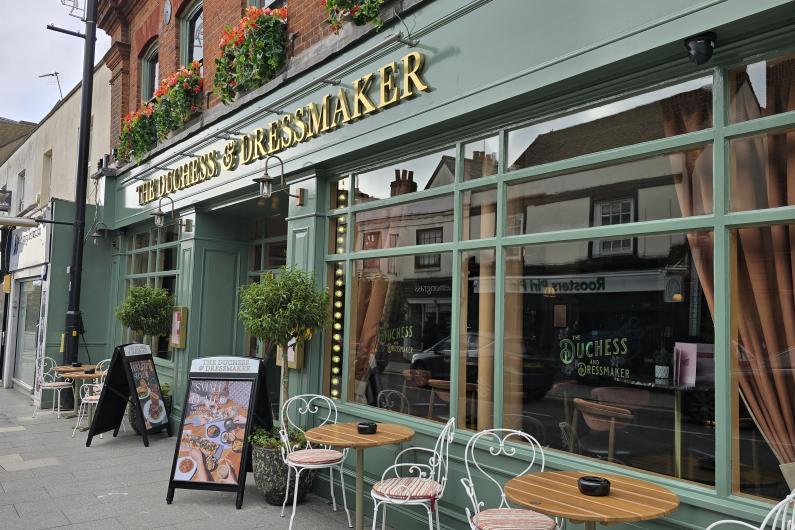 Image showing the exterior of The Duchess & Dressmaker, painted green with gold writing and velvet curtains at the windows.  Two tables and four chairs are outside, while window boxes with red flowers are visible outside the first floor windows.