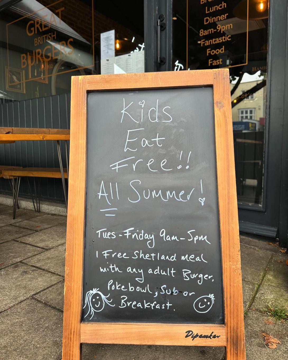 Chalkboard in a frame, positioned outside Hamburger by Hepburns in Shenfield.  Writing on the board reads 'Kids Eat Free!!  All Summer!  Tues - Friday 9am - 5pm   1 Free Shetland meal with any adult burger, Poke bowl, sub or breakfast