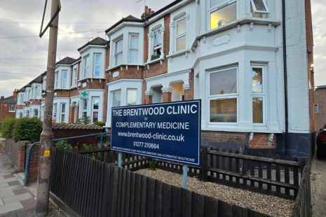 The image shows the exterior of a building identified as "The Brentwood Clinic of Complementary Medicine." The clinic is located in a row of traditional, two-storey, red-brick terraced houses with white window frames and blue-painted accents. A prominent sign in the front yard reads:  "THE BRENTWOOD CLINIC OF COMPLEMENTARY MEDICINE" "www.brentwood-clinic.co.uk" "01277 210664"  The sign is blue with white lettering and stands in a garden area enclosed by a low black wooden fence. 