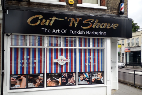 Front view of Cut "N" Shave' barber shop with a sign that reads 'The Art of Turkish Barbering'. The shop has a black awning with gold and white lettering. The windows are decorated with red, white, and blue vertical stripes, resembling a traditional barber's pole. Several photographs on the window show men receiving haircuts and shaves. There are also signs on the window indicating that gift vouchers are available and that walk-in service is offered. A barber pole is visible on the wall.