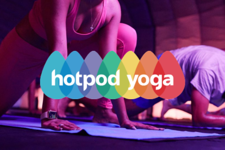 Two people practising yoga inside a dark, softly lit space. The person in the foreground is wearing pink athletic wear, in a low lunge position on a yoga mat. A colourful, egg-shaped 'Hotpod Yoga' logo is centred over the image.