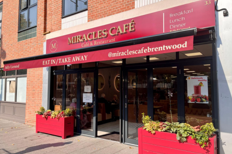 Image of the front of MIRACLES CAFE in Brentwood High Street.  The awning and signage is red with gold text, while two slatted planters are outside on each side of the door.