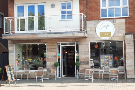 Image showing the exterior of Café  25a in Brentwood.  The ground floor of a building is panelled with light coloured wood, while a circular white sign with gold text shows the words 'Café  25a'.  There are four silver tables with 11 chairs around them at the front of the café .